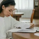 Seorang perempuan sedang membaca berkas di atas meja dengan laptop yang menyala