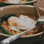 Potret semangkuk mie kuah dengan sayur dan sendok serta sumpit ditata di atasnya.