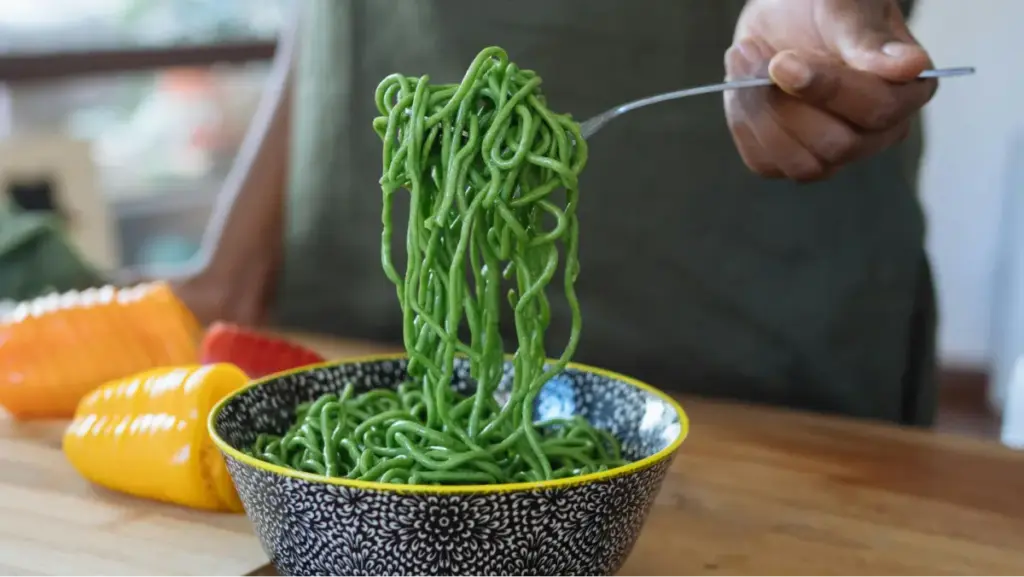 Potret seseorang mengambil mie berwarna hijau menggunakan garpu dari mangkuk bermotif