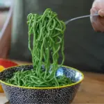 Potret seseorang mengambil mie berwarna hijau menggunakan garpu dari mangkuk bermotif