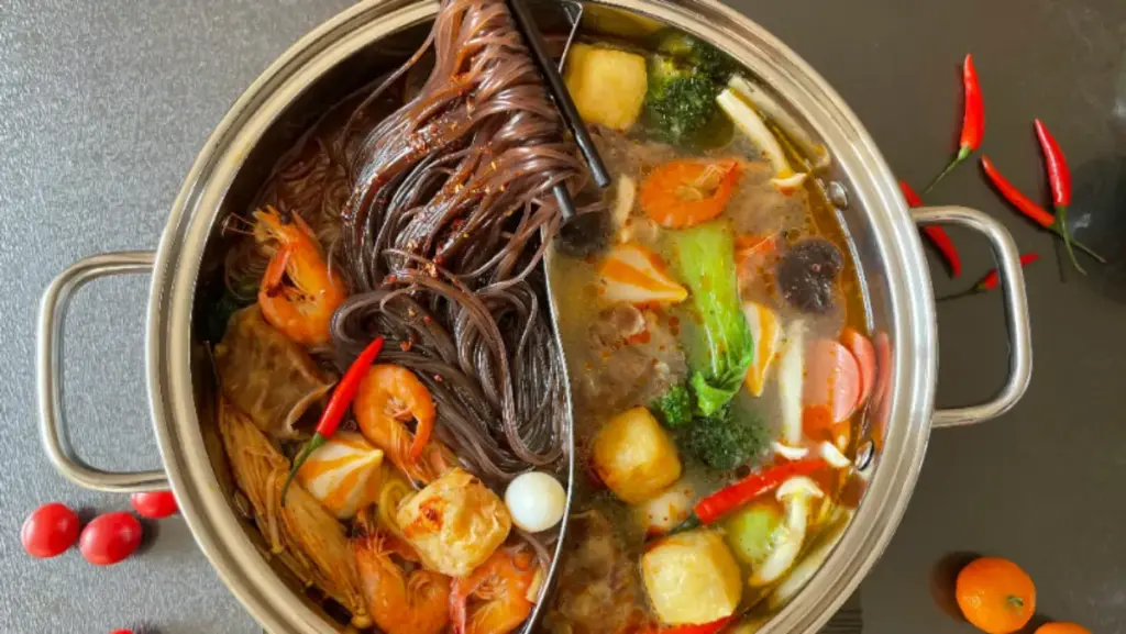 Potret panci dengan sekat berisikan kuah, sayur, seafood, dan mie yang diangkat menggunakan sumpit.