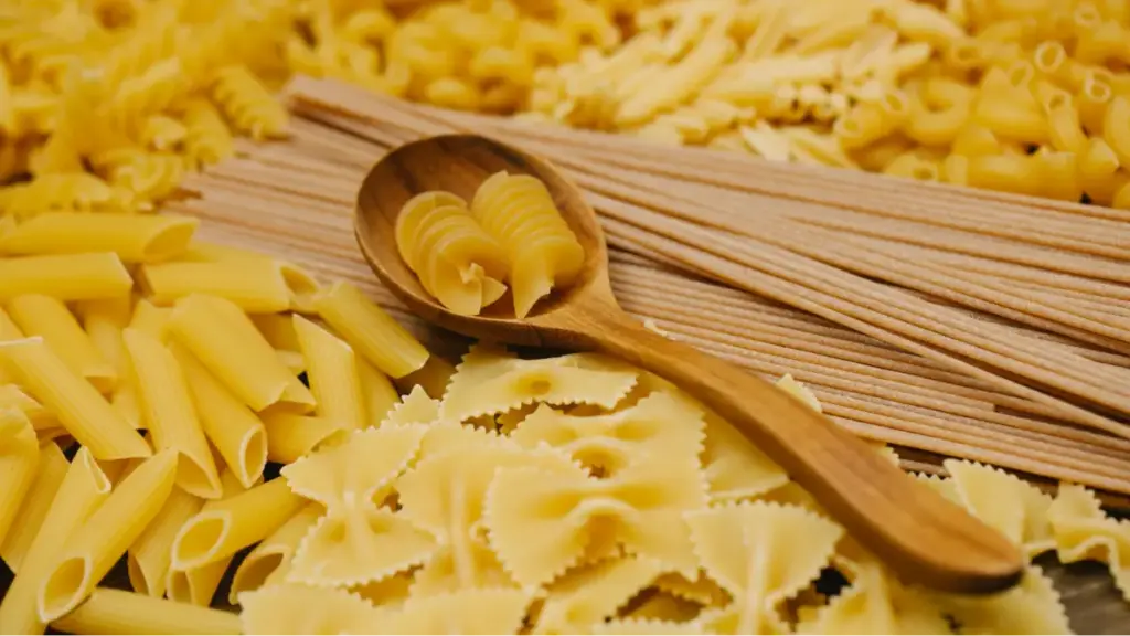 Deretan berbagi macam jenis pasta ditata bersebelahan dengan pasta fusili di atas sendok kayu.
