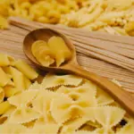 Deretan berbagi macam jenis pasta ditata bersebelahan dengan pasta fusili di atas sendok kayu.
