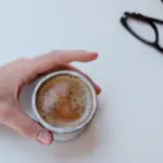 Potret seseorang memegang segelas kopi dnegan kacamata tergeletak di dekatnya