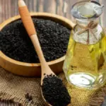 Potret sepiring biji wijen hitam dan sendok berisikan wijen hitam ditata bersebelahan dengan sebotol minyak.