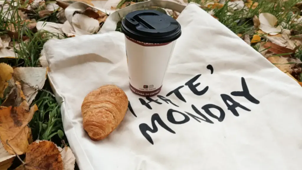 Potret totebag bertuliskan I Hate Monday tergeltak di atas rerumputan dengan secangkir kopi dan croissant