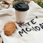 Potret totebag bertuliskan I Hate Monday tergeltak di atas rerumputan dengan secangkir kopi dan croissant