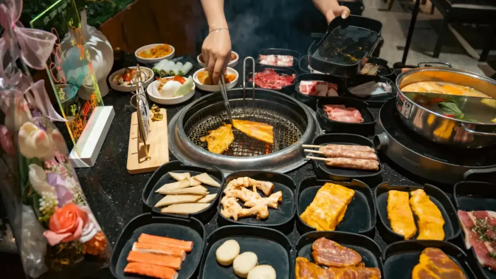 Potret seseorang memanggang daging di meja all you can eat dengan berbagai bahan makanan